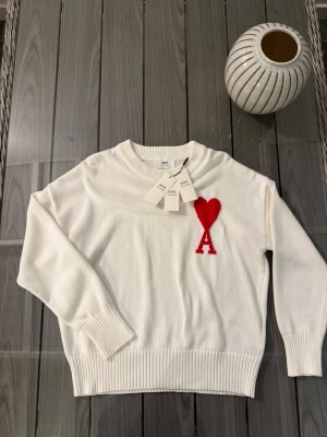 Vit stickad tröja från Ami Paris - Säljer en vit stickad tröja från Ami Paris med röd hjärtlogga och bokstaven A på bröstet. Tröjan har rund hals, ribbade muddar och är långärmad. Perfekt för dig som gillar stilrena plagg med en twist.