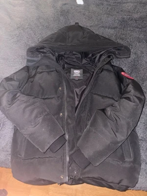 Canada Goose vinter jacka - Svart dunjacka från Canada Goose med huva och klassisk röd logotyp på ärmen. Jackan har dragkedja och tryckknappar framtill, ribbade muddar vid ärmslut och två sidofickor. Perfekt för kalla vinterdagar och har en clean, stilren look.