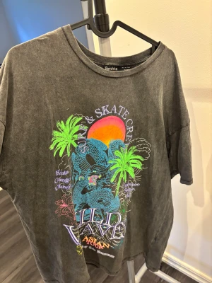 Oversized grå t-shirt Bershka med tryck - Cool oversized grå t-shirt från Bershka med färgglatt tryck av palmer, drake och solnedgång. T-shirten har korta ärmar och en avslappnad passform. Perfekt för dig som gillar streetstyle och unika prints.