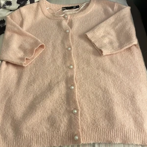 Ljusrosa kofta med pärlknappar - Superfin ljusrosa kofta  med korta ärmar och pärlknappar framtill. Mjuk stickad känsla och ribbade muddar vid ärmslut och nederkant. Perfekt för dig som gillar söta detaljer och vill ha en cozy look.