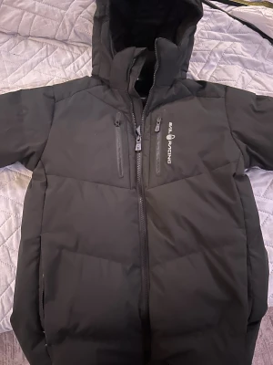 Svart Sail Racing dunjacka med huva - Svart dunjacka från Sail Racing med huva och två bröstfickor med dragkedja. Jackan har Sail Racing-logga på bröstet och patch på ärmen. Snygg, stilren design och varm fyllning, perfekt för kalla dagar. Jackan har hel dragkedja framtill och är riktigt skön att bära.