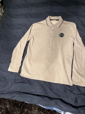 Grå långärmad pikétröja från BOSS - Snygg grå långärmad pikétröja från BOSS med klassisk krage och svarta ränder på kragen. Tröjan har knappar framtill och en rund BOSS-logga på bröstet. Perfekt för dig som gillar stilrena och enkla plagg med premiumkänsla.