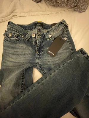 Oanvända True Religion blå bootcut jeans W26 - HELT NYA snygga blå jeans från True Religion med bootcut passform och klassiska kontrastsömmar. Modellen heter Becca och har detaljerade fickor med lock och silverfärgade knappar. Jeansen har låg midja och är tillverkade i slitstarkt denim med gul söm.