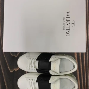 Valentino Garavani svartvita sneakers - Säljer ett par snygga vita Valentino Garavani sneakers med bred svart läderdetalj över vristen och coola nitar på sulan. Skorna har rund tå, platt sula och snörning framtill. Perfekt för dig som gillar exklusiv och stilren design.