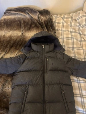 Mörkgrå dunjacka från Polo Ralph Lauren storlek 170/xs - Snygg mörkgrå dunjacka från Polo Ralph Lauren med huva och hög krage. Jackan har flera praktiska fickor med dragkedja och ett diskret Polo-logo på bröstet. Perfekt för kalla dagar med sin puffiga design och stilrena look.