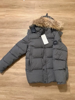 Grå pufferjacka med fuskpäls - Snygg grå pufferjacka från Canada Goose med avtagbar huva och fuskpäls. Jackan har svarta knappar, dragkedja och ett klassiskt märke på ärmen. Perfekt för kalla dagar med sin tjocka vaddering och långa ärmar.