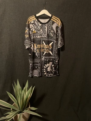 Adidas Real Madrid Bellingham tröja 176 - Säljer en limited edition Real Madrid tröja från Adidas med Bellingham-tryck. Tröjan har svart och guld paisley-mönster, coola grafiska detaljer och guldiga Adidas-ränder på axlarna. Materialet är lätt polyester och perfekt för fotboll eller att rocka på stan.