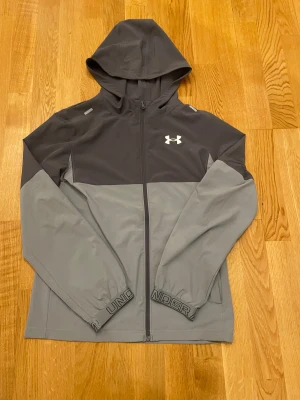 Grå vindjacka från Under Armour - Snygg tvåfärgad vindjacka från Under Armour i mörkgrått och ljusgrått. Jackan har huva, hel dragkedja och logga på bröstet. Elastiska muddar med tryckt text och sportig look. Perfekt för dig som gillar stilren och funktionell design.