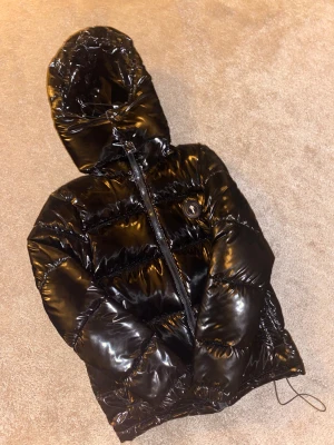 Svart pufferjacka från Trapstar - Säljer en svart, glansig pufferjacka från Trapstar med huva och fet logga på bröstet. Jackan har dragkedja framtill och är riktigt puffig med snygg streetstil. Perfekt för kalla dagar när du vill se fresh ut.