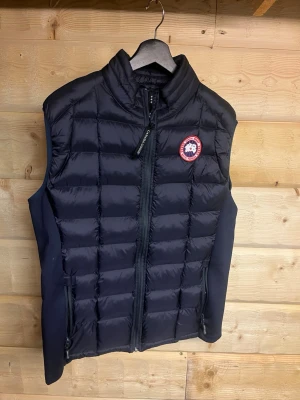 Svart dunväst från Canada Goose L - Svart dunväst från Canada Goose med quiltad design och hög krage. Västen har dragkedja framtill, två sidofickor med dragkedja och klassisk logotyp på bröstet. Materialet är polyester och dun, perfekt för kyliga dagar. Snygg och sportig look med diskreta reflexdetaljer på nacken.