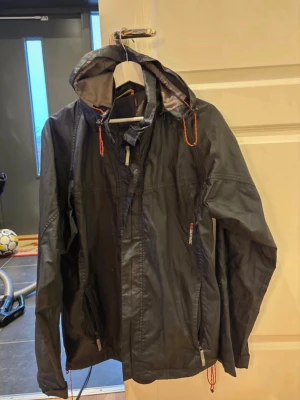Svart vindjacka från Didriksons L - Svart vindjacka från Didriksons i storlek L. Jackan har huva med orange snörning, dragkedja framtill och två fickor med dragkedja. Tillverkad i lätt och slitstark polyester, perfekt för blåsiga dagar. Snygg och funktionell design med diskreta detaljer. Bra skick men snörena ( se bild) är lite slitna. Eller de kanske ska se ut så☺️ vet ej.