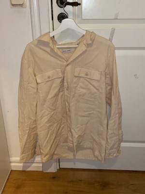 Beige skjorta från Jack & Jones M - Snygg beige skjorta från Jack & Jones i storlek M. Skjortan har två bröstfickor med knappar och klassisk krage. Tillverkad i mjuk bomull, perfekt för en chill och stilren look. Långärmad och med normal passform.