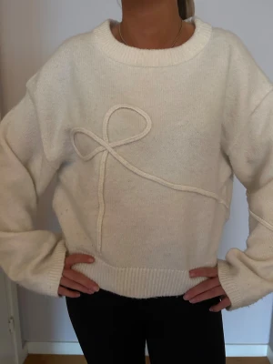 Vit stickad tröja med rosettdetalj - Supermysig vit stickad tröja med  långärmade ärmar. Framsidan har en unik rosettdetalj som ger en personlig touch. Tröjan har breda ribbade muddar vid hals, ärmslut och nederkant. Perfekt för att hålla sig varm och snygg under kyliga dagar. 💓