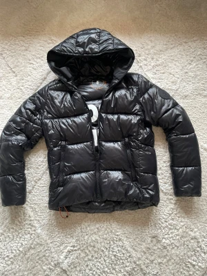 Svart pufferjacka från Save The Duck - Säljer en svart pufferjacka från Save The Duck med huva och två dragkedjeförsedda fickor. Jackan är quiltad och tillverkad i syntetmaterial, helt animal free. Märkets logga finns på ärmen och insidan har tryck med texten '100% Animal Free'. Perfekt för kalla dagar.