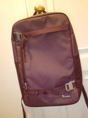 Brun ryggsäck från Db - Säljer en stilren vinröd  ryggsäck från Db med rektangulär form och smarta fack. Väskan har metallspännen, vadderad rygg och axelband samt flera dragkedjor för enkel organisering. Perfekt för dig som vill ha en clean och funktionell look.