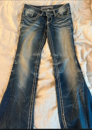 Blå bootcut jeans  - Snygga blå jeans med låg midja och bootcut-ben. Jeansen har kraftiga vita kontrastsömmar, slitningar och detaljerade bakfickor med broderade mönster. Klassisk jeansknapp och läderpatch med logga baktill. Perfekt för dig som gillar statement-jeans.