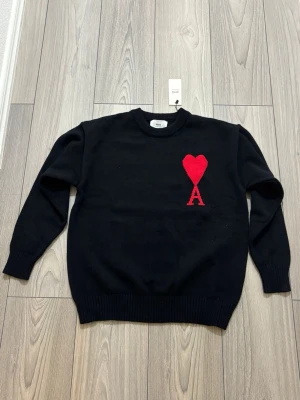 Svart stickad tröja från Ami Paris - Svart stickad tröja från Ami Paris med rund halsringning och ribbade muddar. På bröstet finns ett rött hjärta med bokstaven A, vilket ger en unik och trendig look. Perfekt för dig som gillar stilrena plagg med en twist.