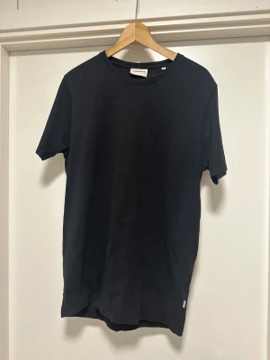 Svart t-shirt från Lindbergh White - En stilren svart t-shirt från Lindbergh White i storlek L. Modellen har rund hals, korta ärmar och en diskret etikett vid nederkanten. T-shirten är gjord i mjuk bomull och har en klassisk, normal passform som funkar till många olika looks.