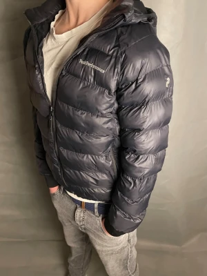 Peak Performance jacka - Snygg svart dunjacka från Peak Performance med huva och dragkedja. Jackan är quiltad och har en sportig look med logga på bröstet och ärmen. Perfekt för kyliga dagar och har en skön, normal passform. Storleken är rekommenderad för 160. Det är tre små defekter som är lagade men de synts knappast.