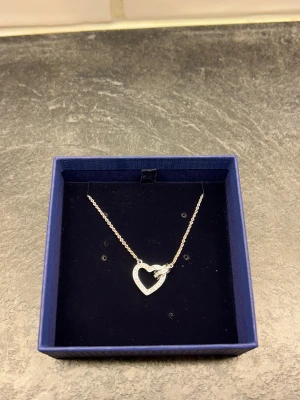 Swarovski silverfärgat hjärthalsband - Snyggt halsband från Swarovski i silverfärg med ett glittrande hjärthänge dekorerat med små kristaller. Kedjan är tunn och hänget har en liten ring som går igenom hjärtat. Helt oanvänt och orört, nypris 1550kr 