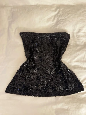Svart glittrig paljett-tubtopp - Säljer en svart tubtopp som är helt täckt med glittriga paljetter. Toppen har en bandeauform och är axelbandslös.