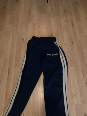 Palm Angels blå mjukisbyxor med vita ränder - Snygga blå mjukisbyxor från Palm Angels med vita sidoremsor och logga på vänster ben. Byxorna har elastisk midja och dragkedja vid benslut för extra stil. Perfekta för en chill och sportig look.