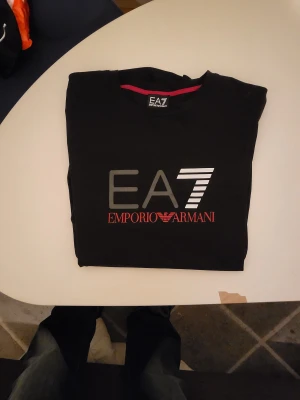 Svart EA7 Emporio Armani t-shirt - Snygg svart t-shirt från EA7 Emporio Armani med stort tryck på bröstet i grått, vitt och rött. Klassisk rund halsringning och stilren design, perfekt för dig som gillar exklusiva märken och streetstyle. I nyskick, storlek L men passar M också då den är mindre i storlek. 