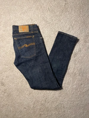 Mörkblå jeans från Nudie Jeans - Snygga mörkblå skinny jeans från Nudie Jeans med klassisk orange söm och vågig broderad detalj på bakfickorna. Modellen har fem fickor, knappgylf och smal passform som sitter tajt längs benen. Perfekt för dig som gillar stilrena och moderna jeans.