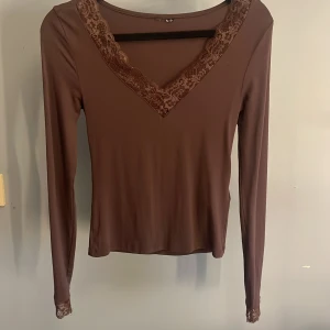 Brun långärmad topp med spetsdetaljer - Säljer en brun långärmad topp med v-ringning och fina spetsdetaljer längs halsringningen och vid ärmsluten. Toppens material är mjukt och stretchigt, vilket gör den bekväm att bära. Perfekt för dig som gillar en feminin och stilren look.
