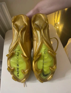Nike Mercurial Elite fotbollsskor guld - Säljer ett par Nike Mercurial fotbollsskor i en riktigt fet guldfärg med stickad ovandel och snörning. Skorna har en platt sula med dobbar för gräsplan och coola detaljer som Mercurial-loggan på sidan. Perfekt för dig som vill sticka ut på planen.