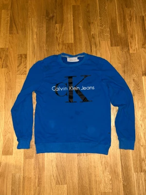 Blå Calvin Klein Jeans tröja S - Snygg blå tröja från Calvin Klein Jeans i storlek S. Tröjan har ett stort CK-logotryck i svart på bröstet och rund hals. Tillverkad i mjuk bomull med ribbade muddar vid ärmslut och nederkant. Perfekt för dig som gillar stilrena och klassiska plagg.