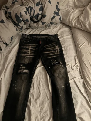 Svarta Dsquared2 jeans med slitningar - Säljer ett par svarta jeans från Dsquared2 med coola slitningar, patchdetaljer och blekta effekter framtill. Jeansen har klassisk femficksdesign, Dsquared2-logga bak och är tillverkade i jeansmaterial med en skinny passform.