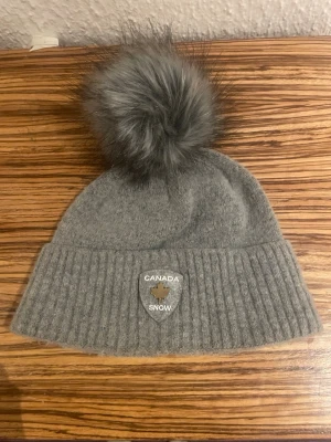 Grå Canada Snow mössa med pälsboll - Säljer en snygg grå mössa från Canada Snow med stor fluffig pälsboll på toppen och ribbad kant. Märket har en silverfärgad logga framtill. Mössan är stickad i mjukt material och passar perfekt för kalla dagar.