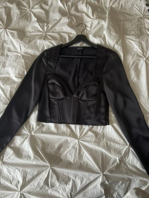 Svart satin korsettopp med långa ärmar - Snygg svart korsettopp från ginatricot i glansigt satinmaterial. Toppen har långa ärmar, markerade kupor och croppad passform som ger en cool siluett. Perfekt för dig som vill ha en trendig och edgy look med lite extra attityd.