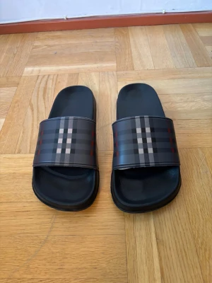 Svarta sandaler med rutigt mönster - Säljer ett par svarta sandaler från Burberry med klassiskt rutigt mönster i grått, beige och rött över remmen. Snygg och stilren design med platt sula och öppen tå. Tillverkade i syntetmaterial, perfekta för sommaren och chill dagar.