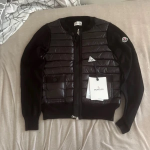 Svart Moncler - Snygg svart jacka från Moncler med quiltad, glansig front och två fickor med knapp. Ärmarna är stickade och har ribbade muddar. Jackan har dragkedja framtill och klassisk Moncler-logga på ena ärmen. Perfekt för dig som vill ha en stilren och exklusiv look.