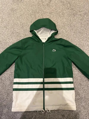 Grön vindjacka från Lacoste Sport - Snygg grön vindjacka från Lacoste Sport med huva och hel dragkedja. Jackan har vita och gröna ränder nertill, meshfoder och stor LACOSTE-logga i gult och vitt på ryggen. Klassisk krokodillogga på bröstet och dragsko nertill för extra passform. 