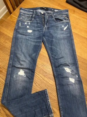Replay Anbass blå jeans, storlek 31/32 - Säljer ett par Replay Anbass jeans i klassisk blå tvätt med slitna detaljer och snygga slitningar på benen. Modellen har raka ben, normal passform och dragkedja i gylfen. Materialet är mjukt jeans och bakfickan har Replay-logga i brunt läder. I nyskick!!