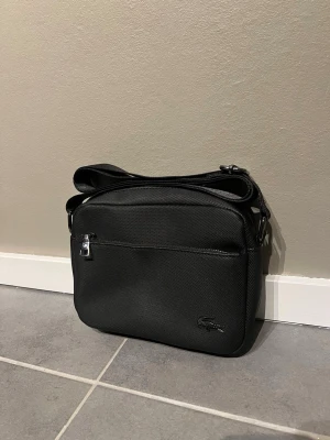 Svart axelväska från Lacoste - Snygg svart axelväska med stilren design och justerbar rem. Väskan har flera fack med dragkedja och är tillverkad i slitstark syntet. Perfekt som julklapp och för att bära dina essentials.