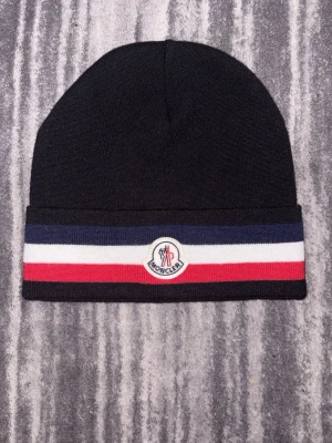 Svart Moncler mössa med ränder - Svart mössa från Moncler med breda ränder i marinblått, vitt och rött längs kanten. Framtill sitter Monclers klassiska logotyp. Mössan är stickad och har en enkel, rundad form som passar perfekt för kalla dagar.
