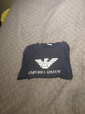 Svart t-shirt från Emporio Armani - Svart t-shirt från Emporio Armani med stort vitt logotryck framtill. Klassisk rund halsringning och korta ärmar. Perfekt för dig som gillar stilrena och ikoniska plagg med tydlig branding.