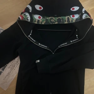 Svart BAPE Shark hoodie med camo-detaljer - Svart hoodie från BAPE med ikonisk shark-hood, camo-mönster på luvan och broderade detaljer i färg. Dragkedja framtill och tryckta loggor längs insidan. Perfekt statement-plagg för streetwear-älskare.