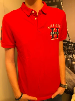 Röd Hilfiger Denim piké med brodyr - Snygg röd pikétröja från Hilfiger Denim med stor broderad logga på bröstet och klassisk krage. Kortärmad modell med knappar framtill och liten flaggdetalj på ärmen. Perfekt för dig som vill ha en sportig och stilren look att använda en skön sommarkväll eller på golfbanan. Knappt använd och inga tecken på slitage. Nypris 599