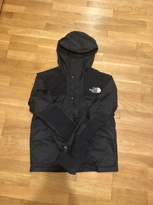 Svart vindjacka från The North Face XS - Svart vindjacka från The North Face i storlek XS. Jackan har huva, långa ärmar och logga på bröstet. Tillverkad i lätt och slitstark polyester med meshfoder för extra komfort. Perfekt för blåsiga dagar och streetwear-vibe.