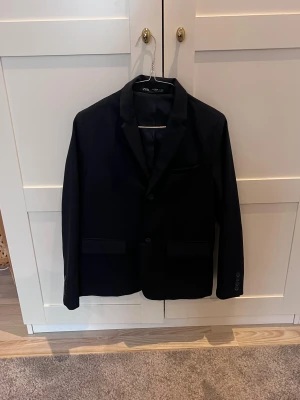 Svart kavaj från Zara - Stilren svart kavaj från Zara med klassisk krage och två knappar framtill. Kavajen har två fickor med lock och en bröstficka, samt knappar vid ärmsluten. Perfekt för en dressad look och passar till många olika outfits.
