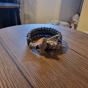 Armband torshammare viking - Säljer ett handgjort armband i brun och beige paracord med en detaljerad Torshammare i metall som lås. Armbandet har grov flätning och coola vikingainspirerade detaljer, perfekt för dig som gillar unika accessoarer. 23,5 cm lång. 3 cm bred.