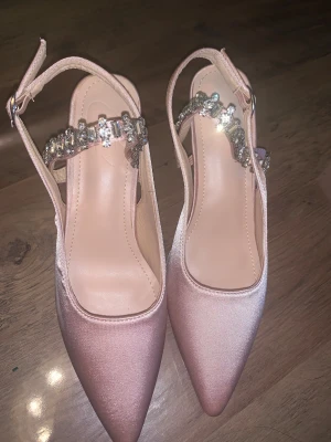 Rosa pumps med strass och klack - Rosa pumps med glittrande strassdetaljer. Justerbart spänne vid ankeln. Oanvända🩷