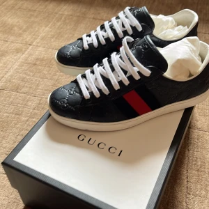 Svarta Gucci sneakers med röd/blå rand - Gucci sneakers i svart skinn med präglat GG-mönster, vita snören och klassiska röda och blå ränder på sidorna. Låg modell med vit sula och rund tå. Snygga detaljer och lyxig känsla – perfekta för dig som vill sticka ut. Eller bara vill vara jävligt grich. Skorna kommer med Gucci box och dustbag för vardera skor. Tillkommer även äkthetsintyg.