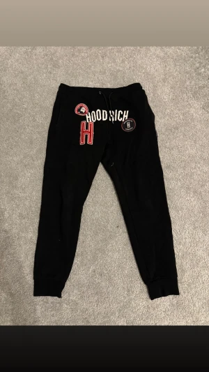 Svarta mjukisbyxor från Hoodrich - Svarta mjukisbyxor från Hoodrich med broderade och tryckta detaljer framtill, inklusive stor röd H och logotyper. Byxorna har snörning i midjan och muddar vid bensluten för en skön och avslappnad passform.