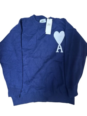 Ami Paris stickad tröja navy - Snygg blå stickad tröja från Ami med rund hals och ribbade muddar. På bröstet finns ett vitt hjärta med bokstaven A, vilket ger en unik och trendig look. Perfekt för dig som gillar stilrena detaljer och vill sticka ut lite extra.